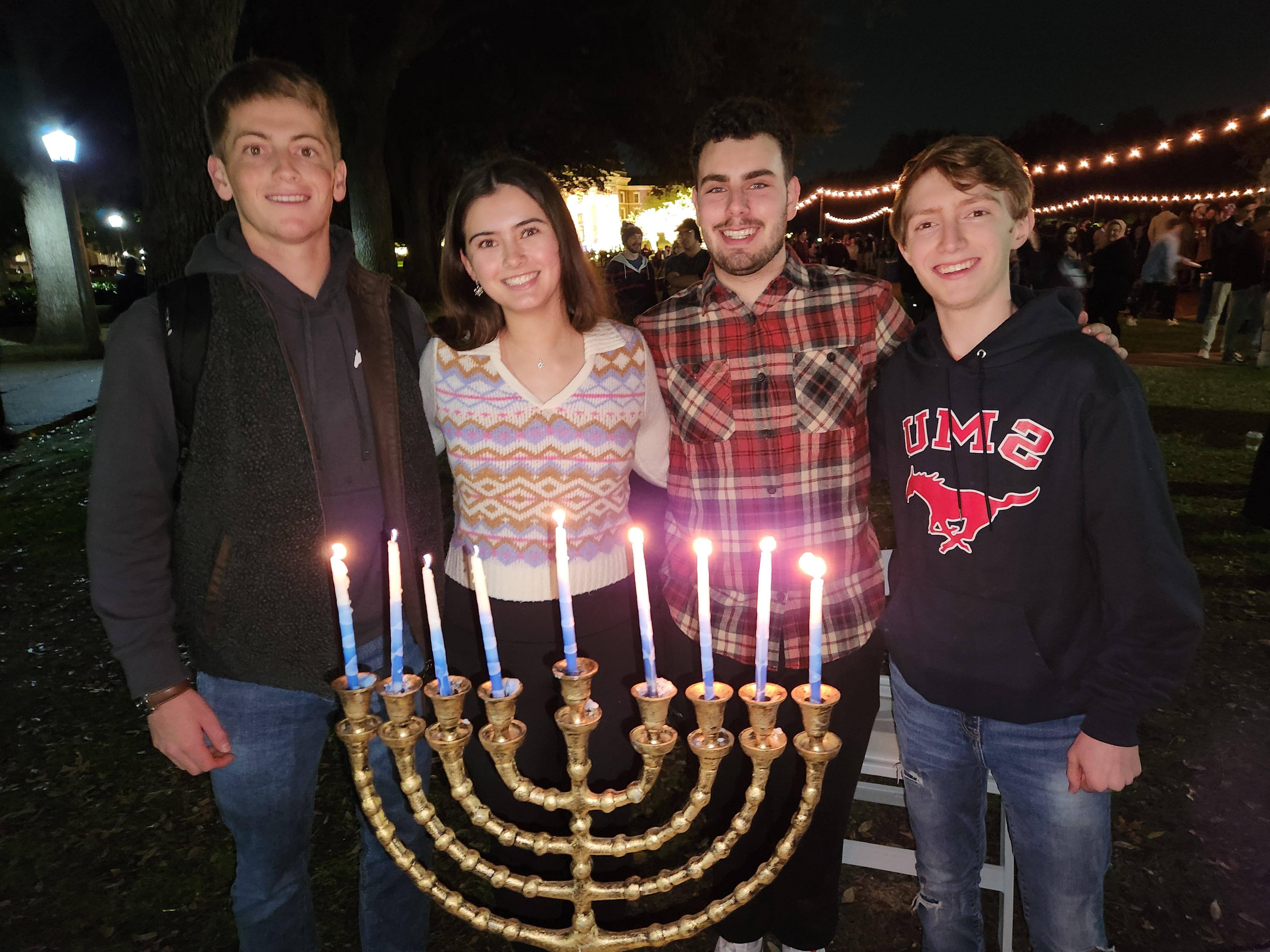 students standing behind a menorah with seven white candles all lit outdoors at night