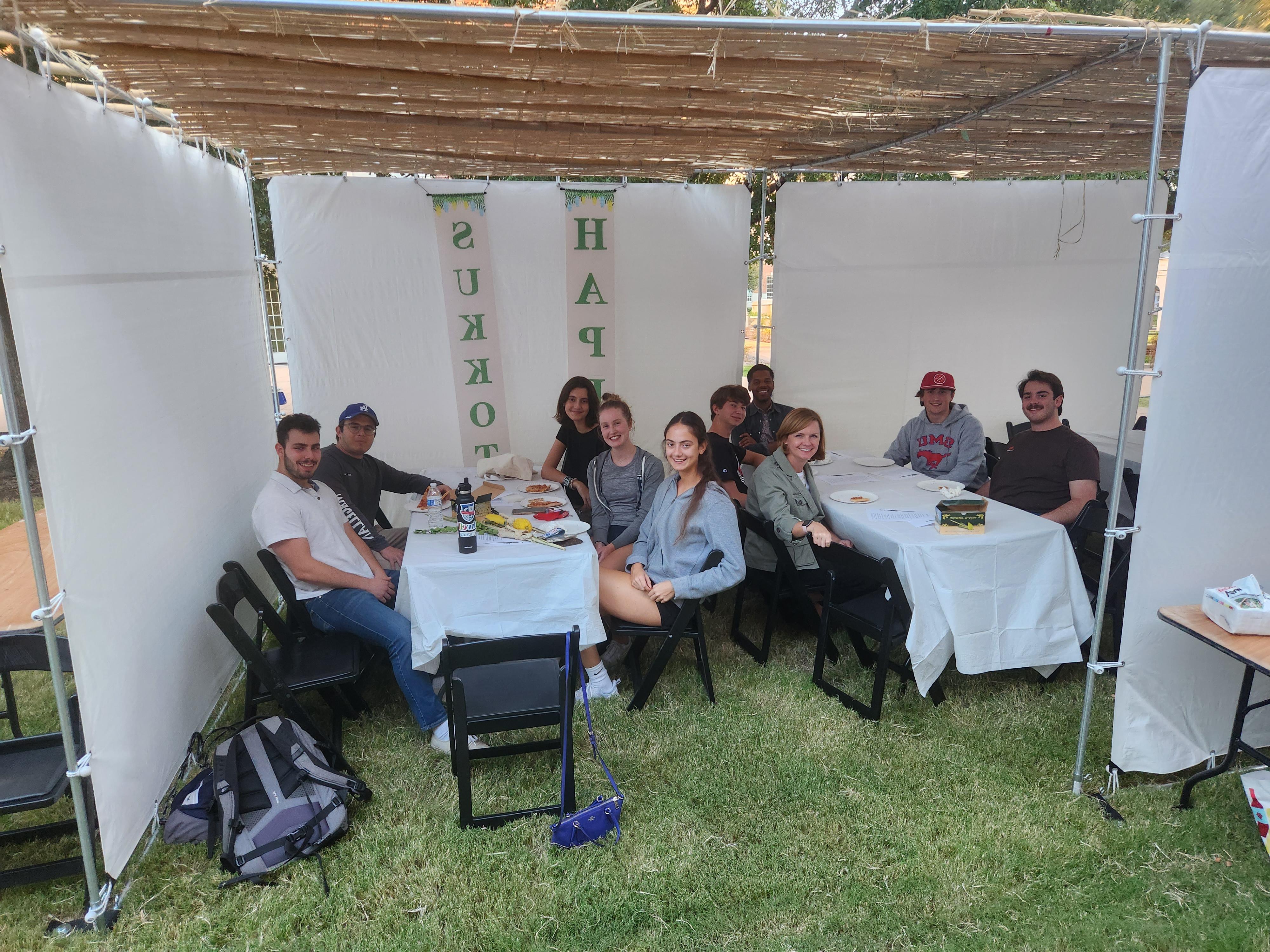 students at tables outdoors on grass surrounded by white drapes covered with string lights